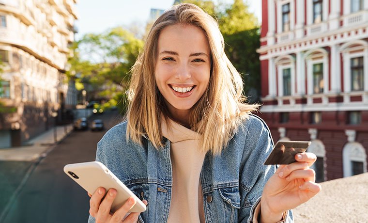 Cheerful young blonde girl shopping online with mobile phone and credit card while standing on a street